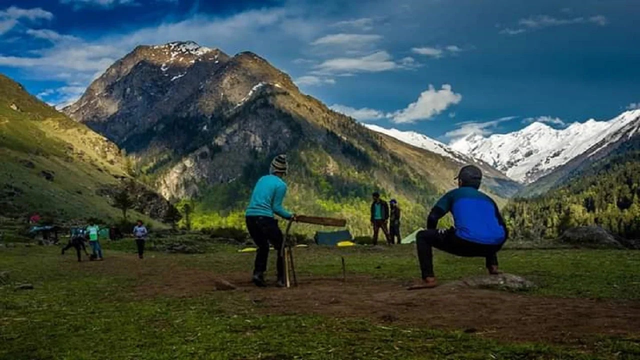 People playing cricket on a lush green field in Har Ki Dun valley, surrounded by majestic Himalayan mountains.