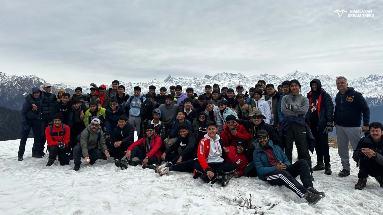 A group of trekkers celebrating at the summit of Dayara Bugyal with stunning Himalayan peaks in the background.